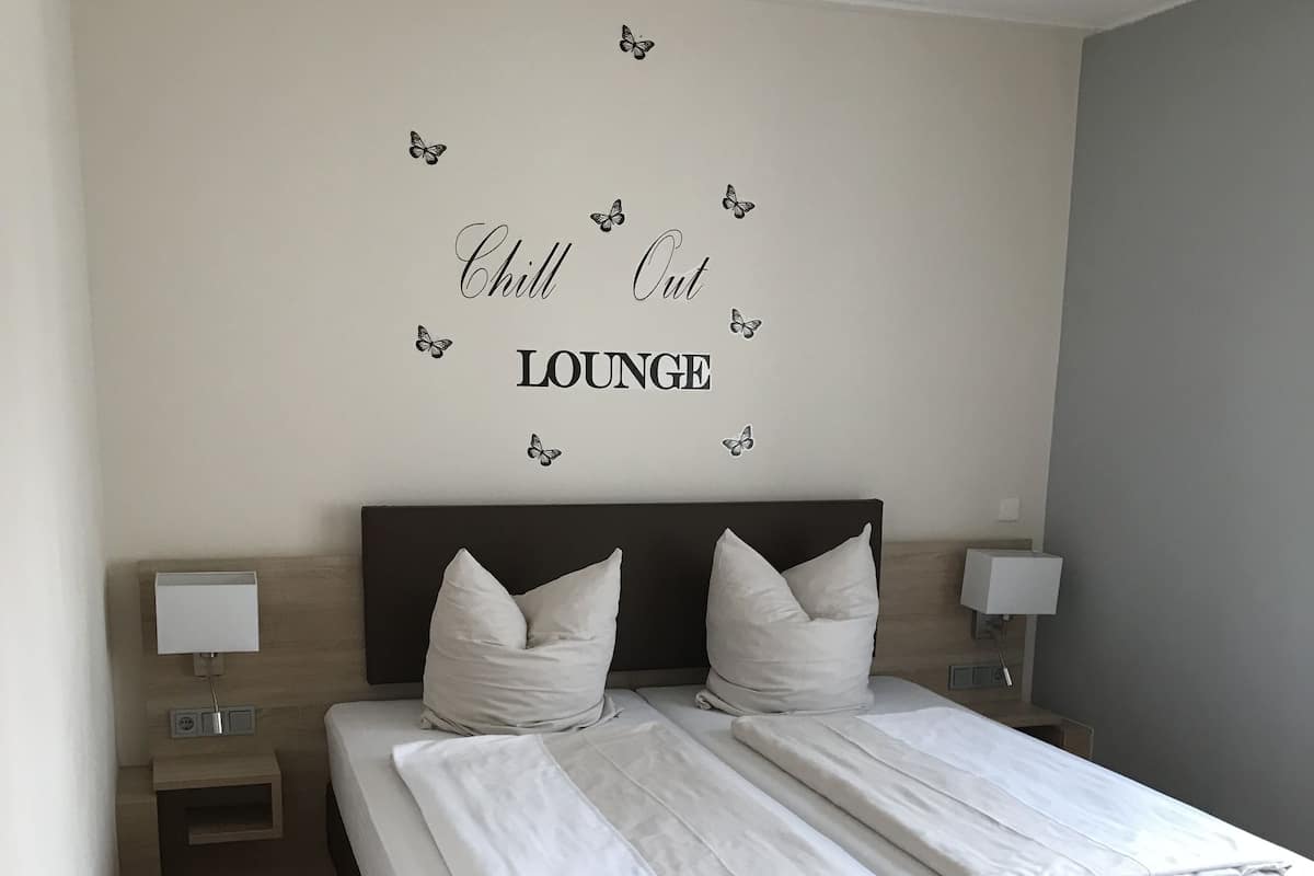 A double bed with white bedding, two nightstands with lamps, and a wall with a 'Chill Out Lounge' sign.