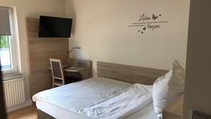 Quarto com cama, escrivaninha, cadeira, televisão fixada na parede e janela.