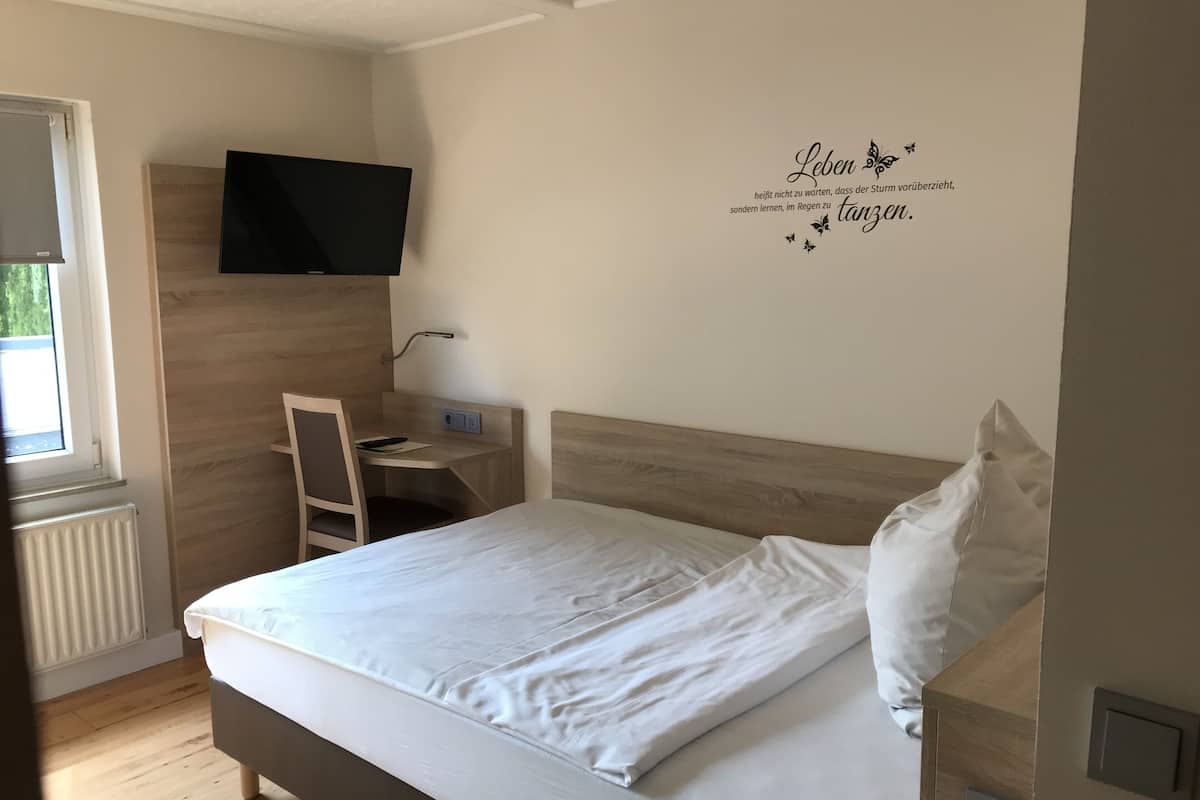 A bedroom with a bed, a desk, a chair, a TV mounted on the wall, and a window.