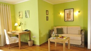 Uma sala de estar com parede verde, sofá bege, mesa de madeira com frutas, luminária e uma cadeira.