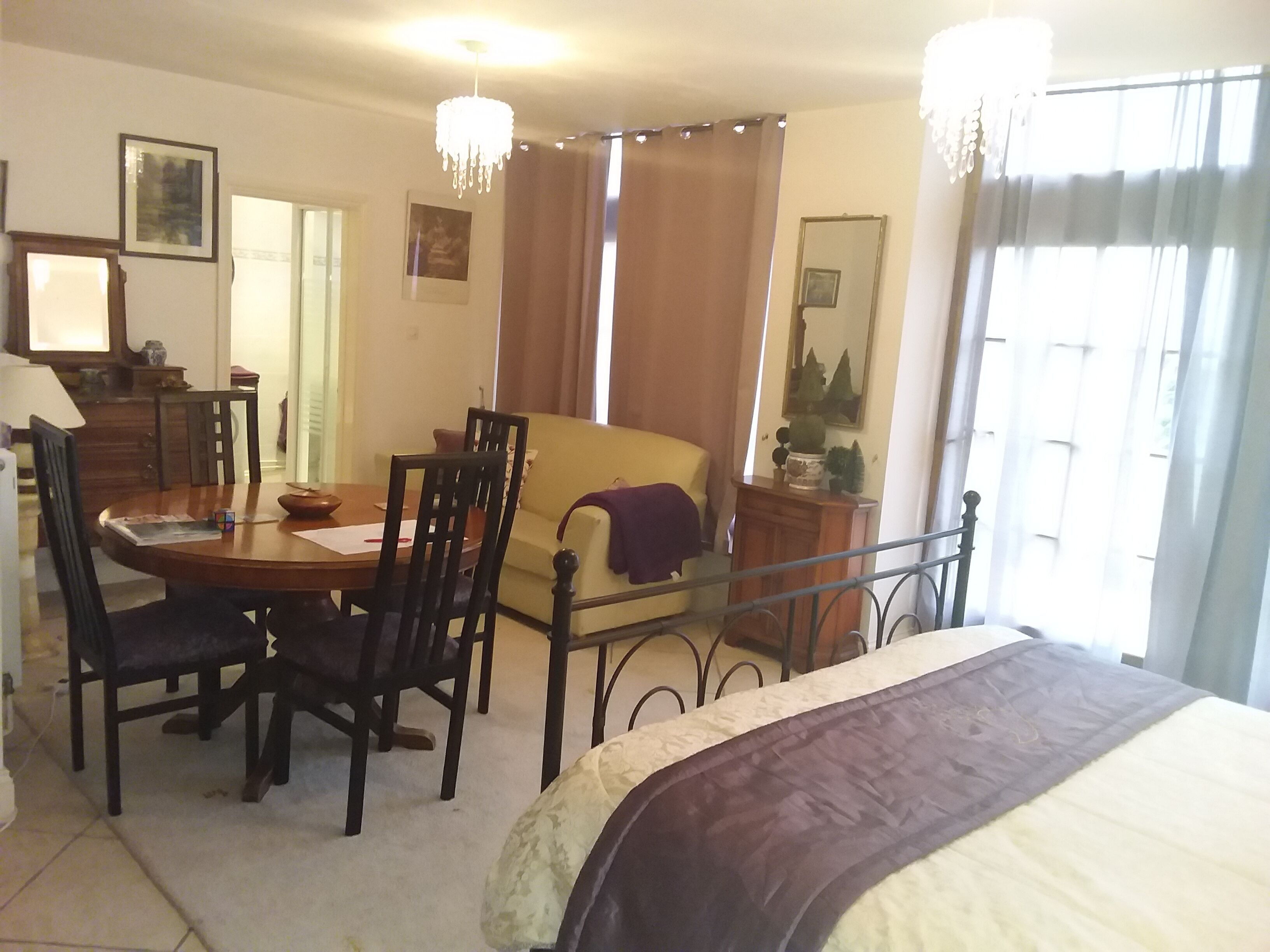 Una habitación de hotel con cama, una mesa de comedor con sillas, un sofá, una araña y una ventana con cortinas.