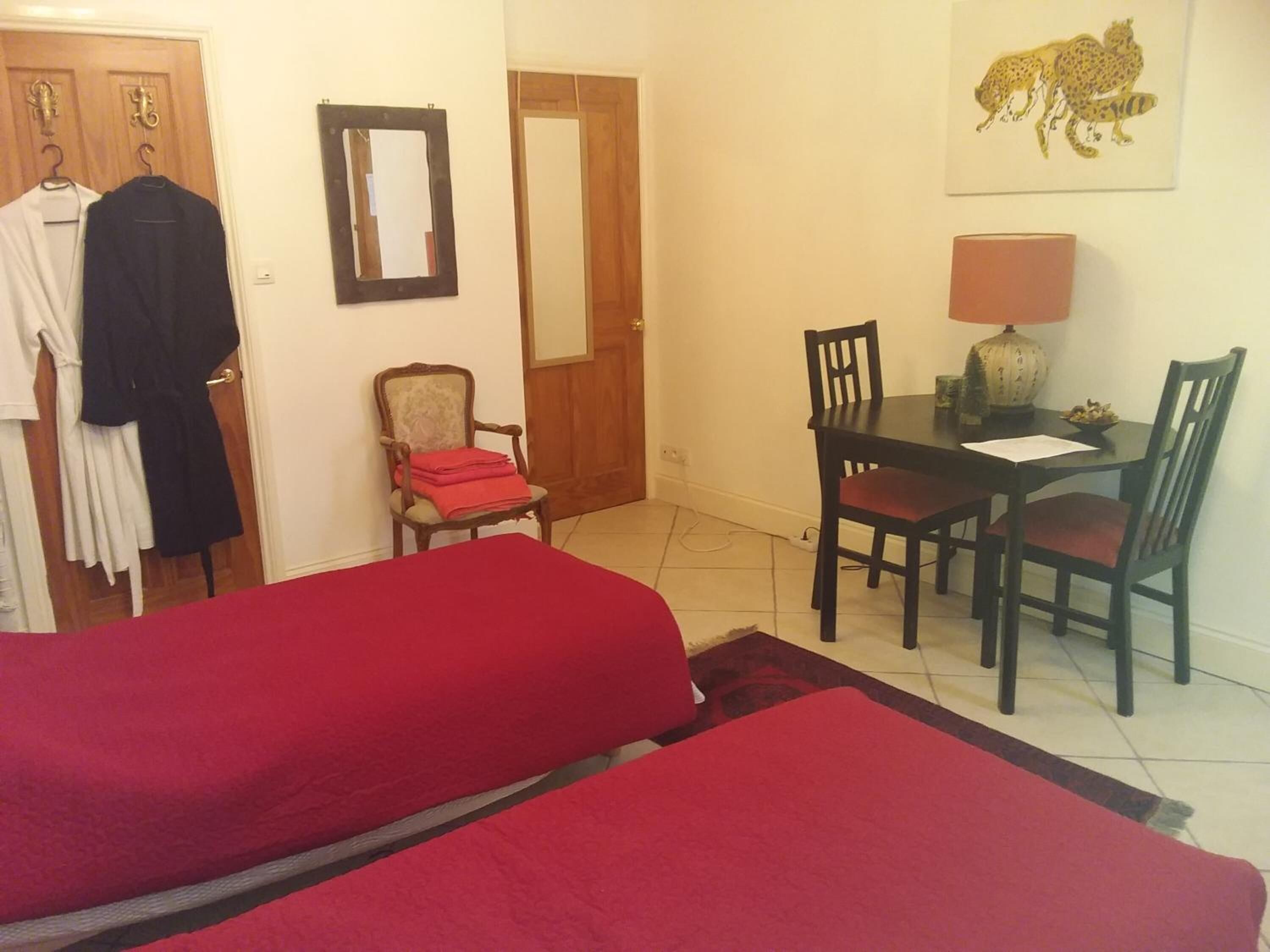 A room with two beds, a dining table with chairs, a chair with a red blanket, a mirror, and a leopard print artwork on the wall.