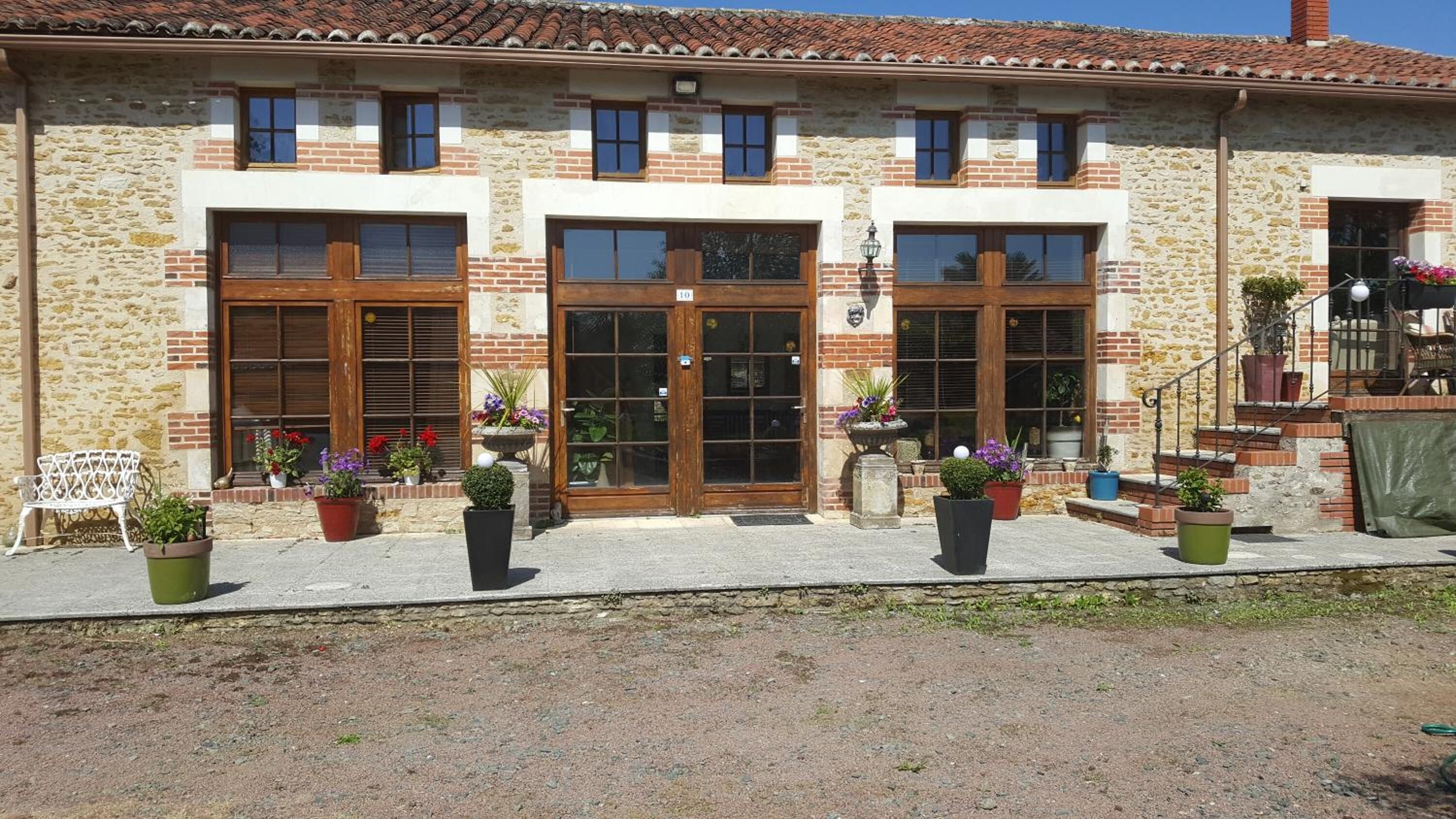 Edifici de pedra de dos pisos amb portes i finestres de fusta grans, una petita zona d'seients a l'exterior amb una boscassa i plantes en testos.