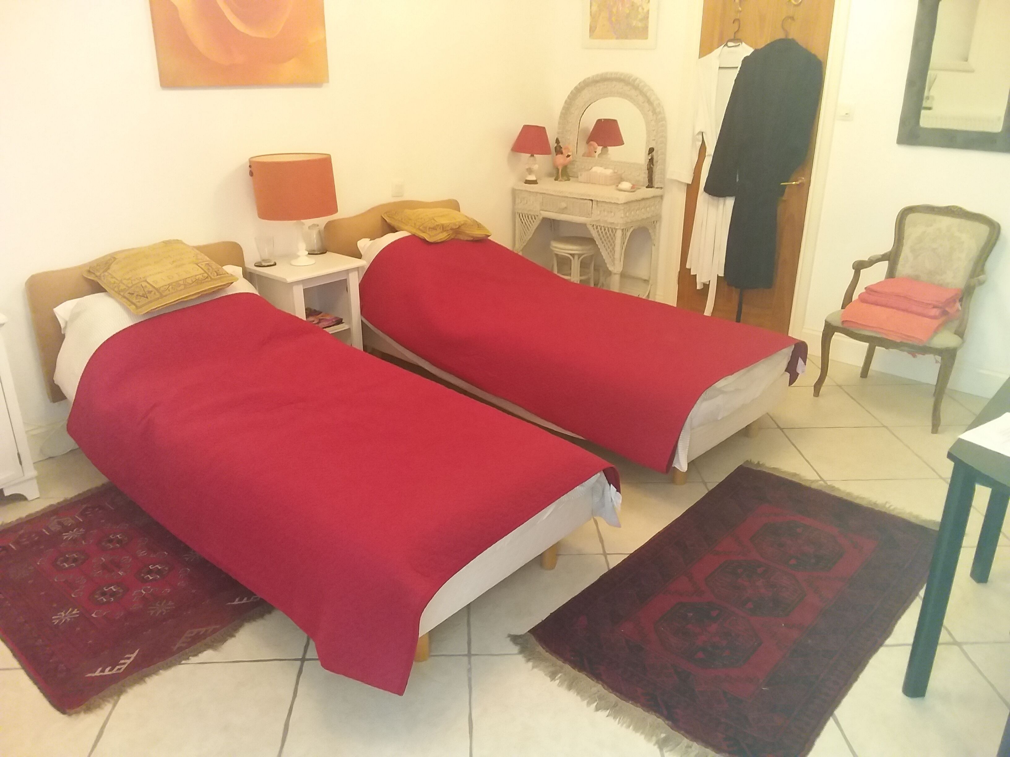 Une chambre avec deux lits simples, une table de chevet avec une lampe, une coiffeuse avec un miroir, une chaise et un manteau accroché.