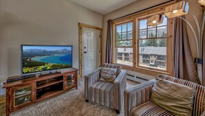 Een woonkamer met een grote flatscreen televisie, een houten tv-meubel, gestreepte banken en een groot raam met uitzicht op gebouwen en bergen.