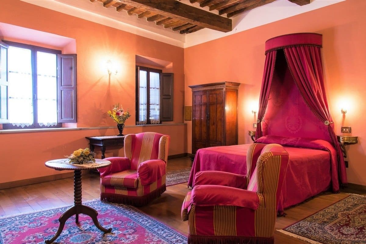 A bedroom with a four-poster bed, a wooden cabinet, a small round table with a vase, and two striped armchairs.