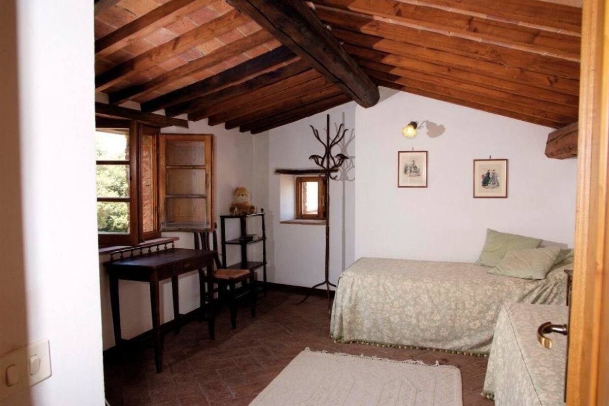 A cozy room with a wooden ceiling, a bed with a floral bedspread, a desk with a chair, and a window with a view of greenery.