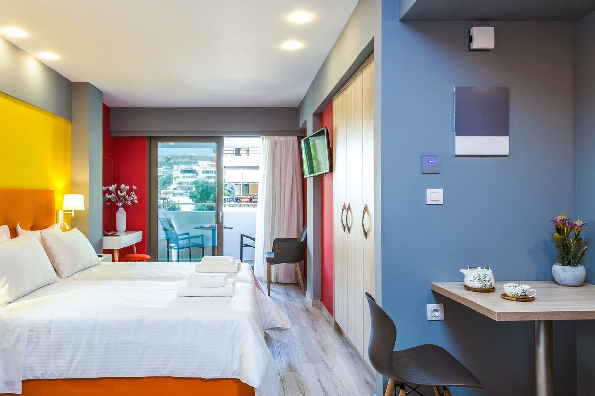 Habitación de hotel con cama, escritorio, silla, televisión y balcón con vista a edificios.