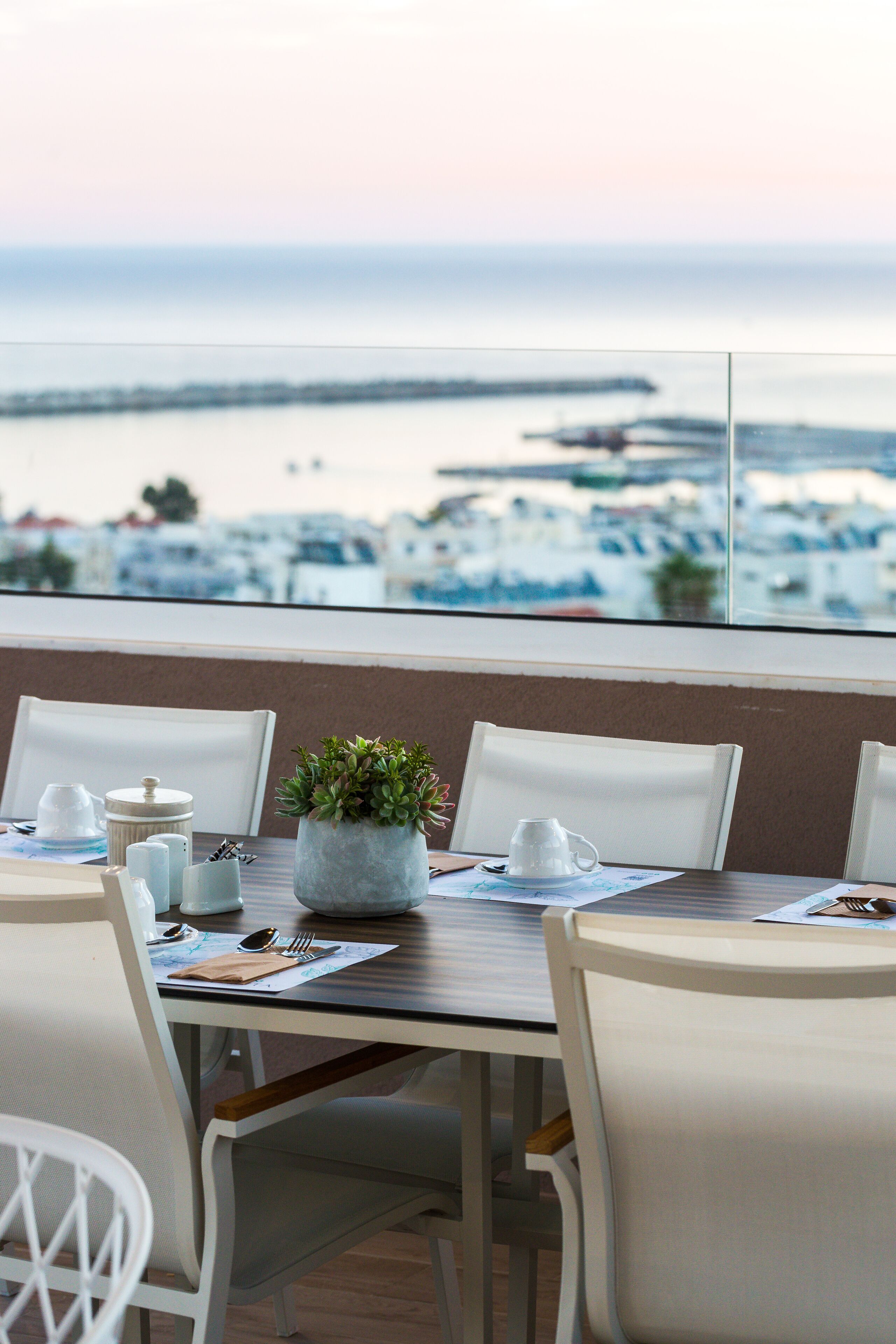 Ein Essbereich mit Blick auf das Meer und einen Steg. Weiße Stühle, ein Tisch mit einer Pflanze und ein Fenster mit Wasserblick.