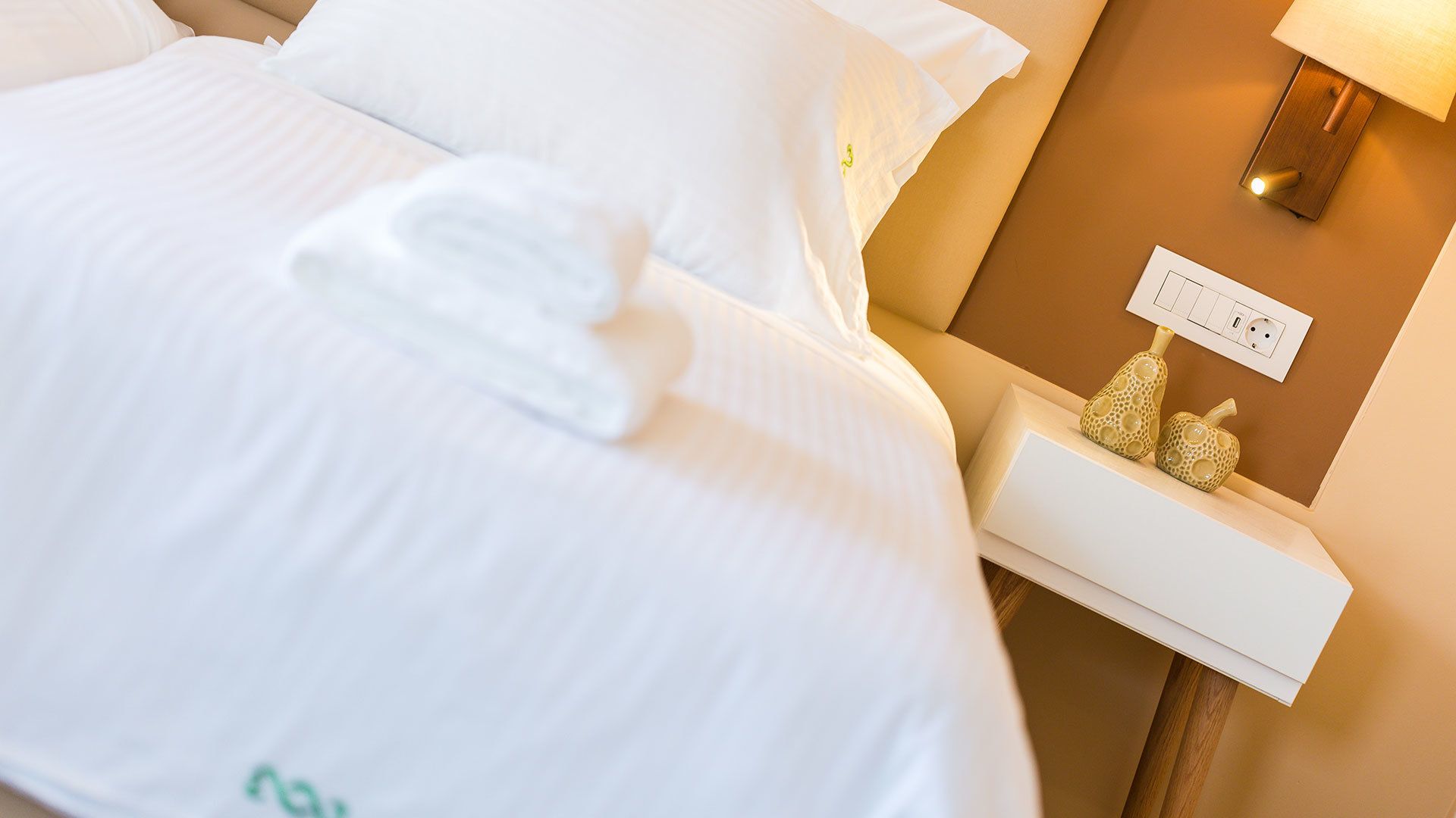 A neatly made bed with white linens and a folded towel, a bedside table with a lamp and decorative items, and a power outlet on the wall.