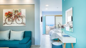 Una habitación de hotel moderna con un sofá color turquesa, un cuadro de una bicicleta, una cama blanca, una mesita con un jarrón y vista al mar a través de la ventana.