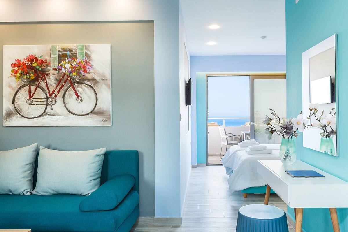 Una habitación de hotel moderna con un sofá color turquesa, un cuadro de una bicicleta, una cama blanca, una mesita con un jarrón y vista al mar a través de la ventana.