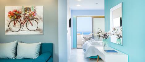 A modern hotel room with a teal sofa, a painting of a bicycle, a white bed, a small table with a vase, and a view of the sea through the window.
