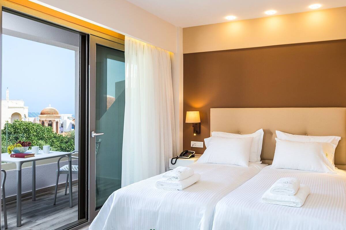 Habitación de hotel con cama, dos mesitas de noche, una lámpara y una ventana con cortinas, y vistas a edificios.