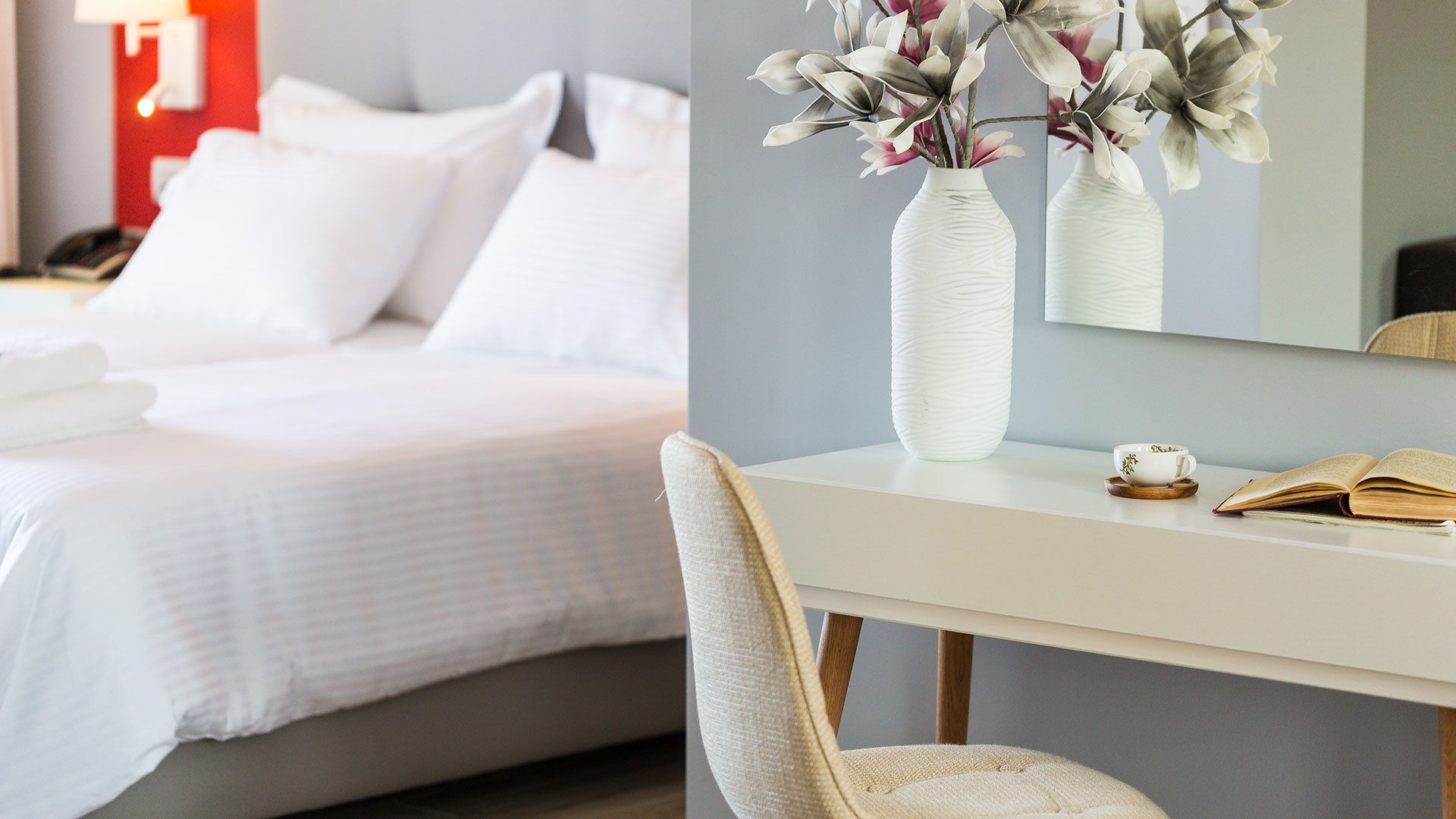 Habitación de hotel con una cama bien hecha, un escritorio blanco con un florero y una silla, además de un libro.