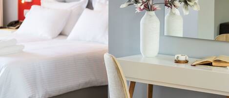 Habitación de hotel con una cama bien hecha, un escritorio blanco con un florero y una silla, además de un libro.