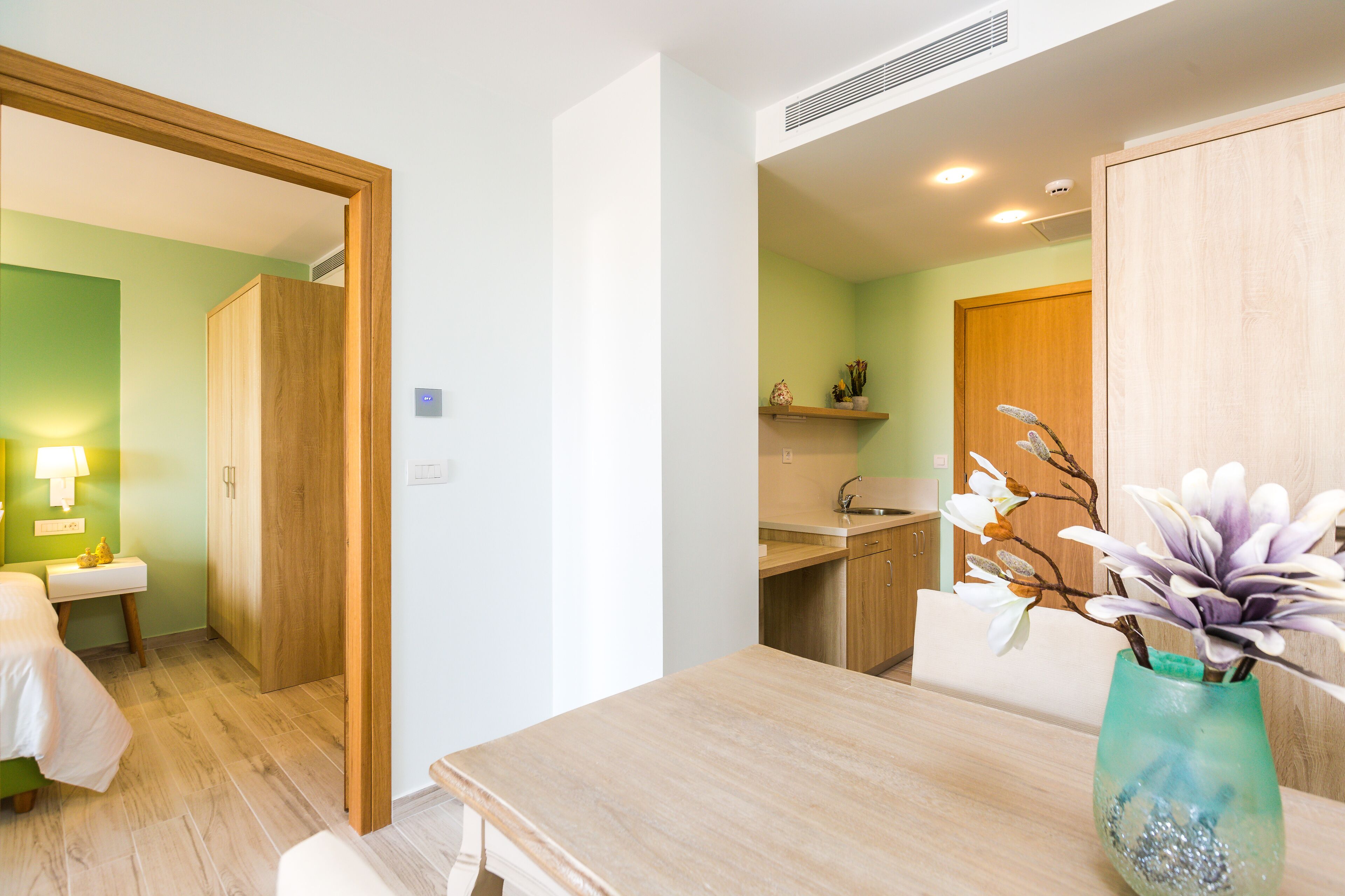 Una habitación de hotel moderna con un armario de madera, un baño pequeño, un rincón de comedor con una mesa, y un jarrón con flores.