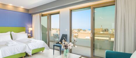 A hotel room with two beds, a sofa, a table with books, a vase with flowers, and a balcony with a view.