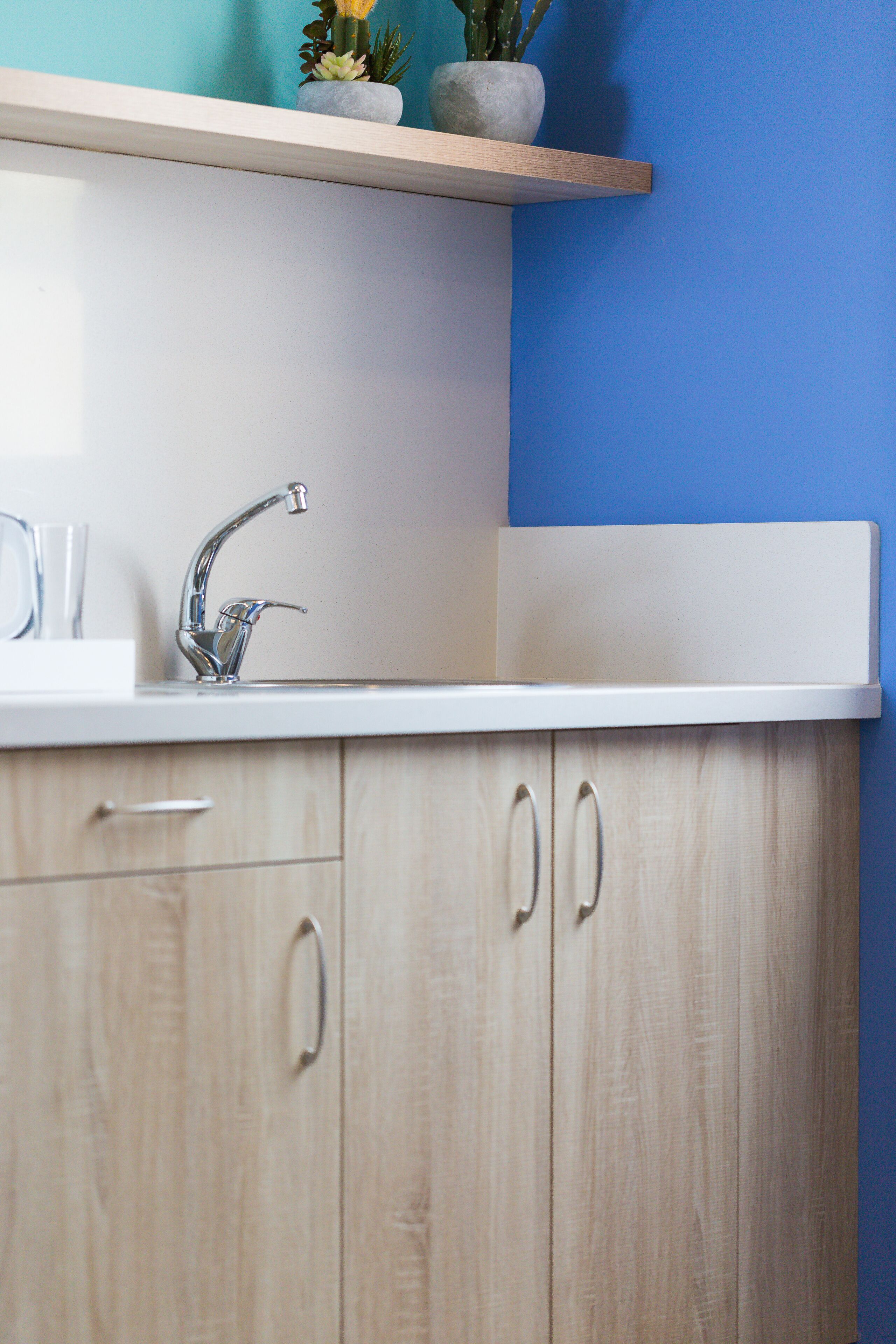 A kitchen with a white countertop, a stainless steel faucet, wooden cabinets, and a blue wall.