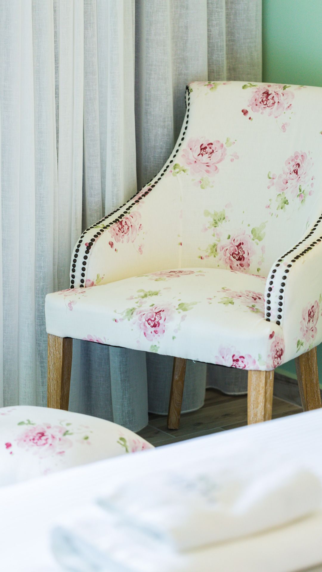 A chair with floral upholstery and wooden legs, positioned in front of a curtain.