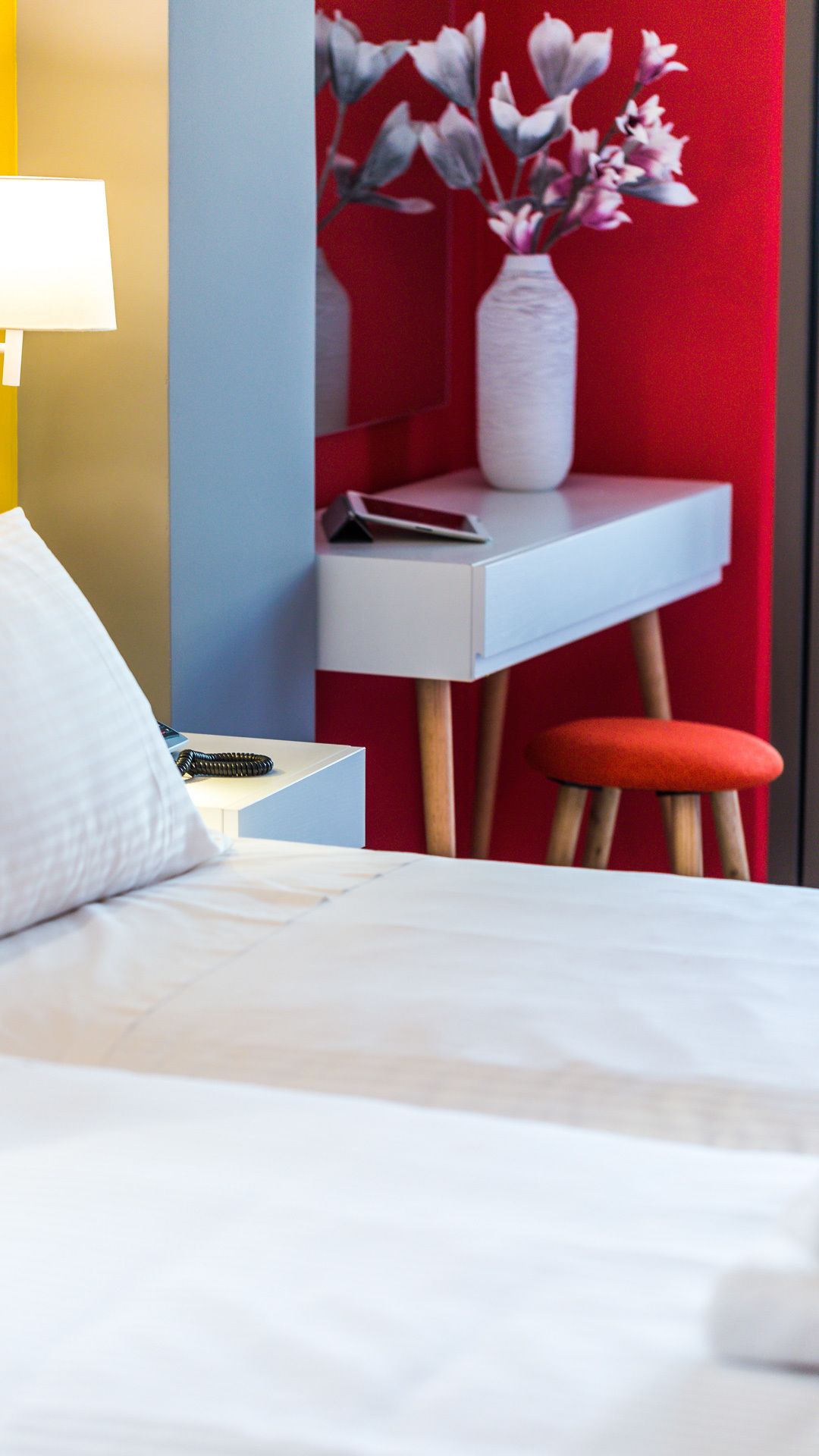 A neatly arranged hotel room with a bed, a desk, a chair, a lamp, and a vase with flowers.