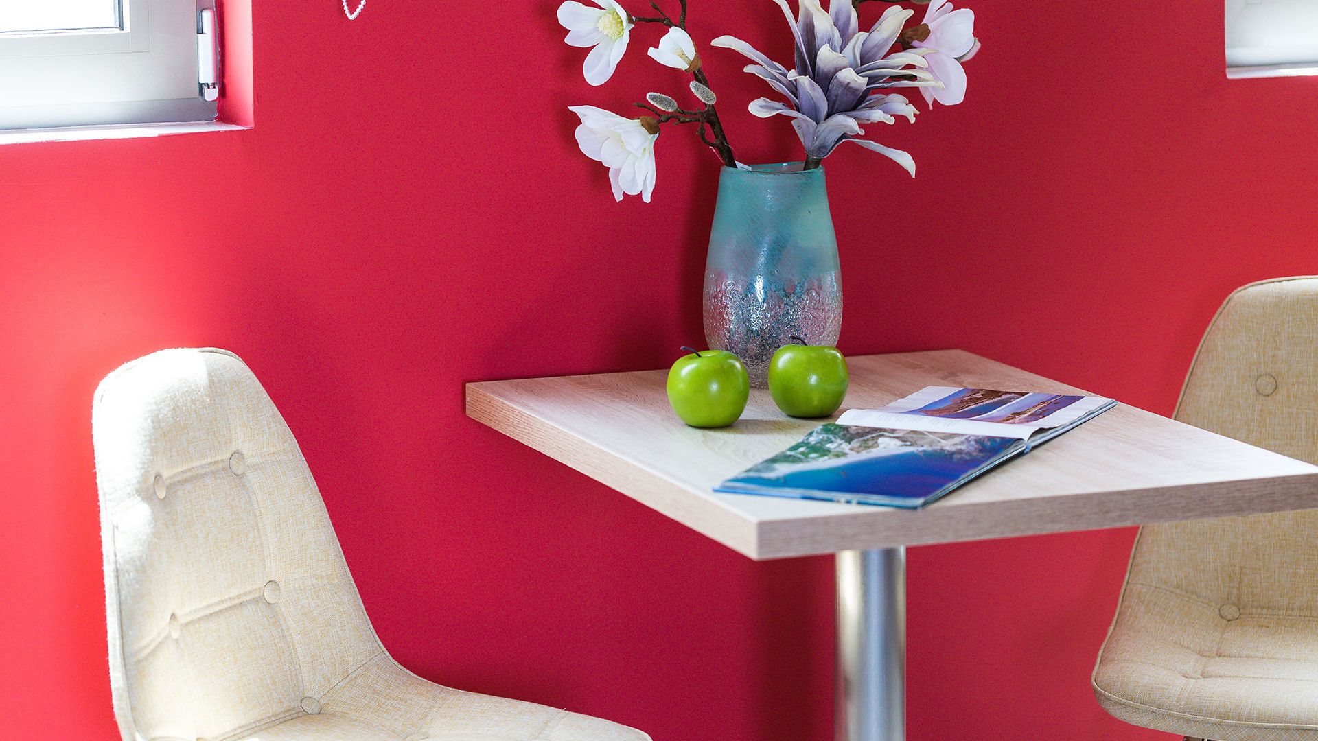 A room with a red wall, a small table with two apples and a vase, and two chairs.