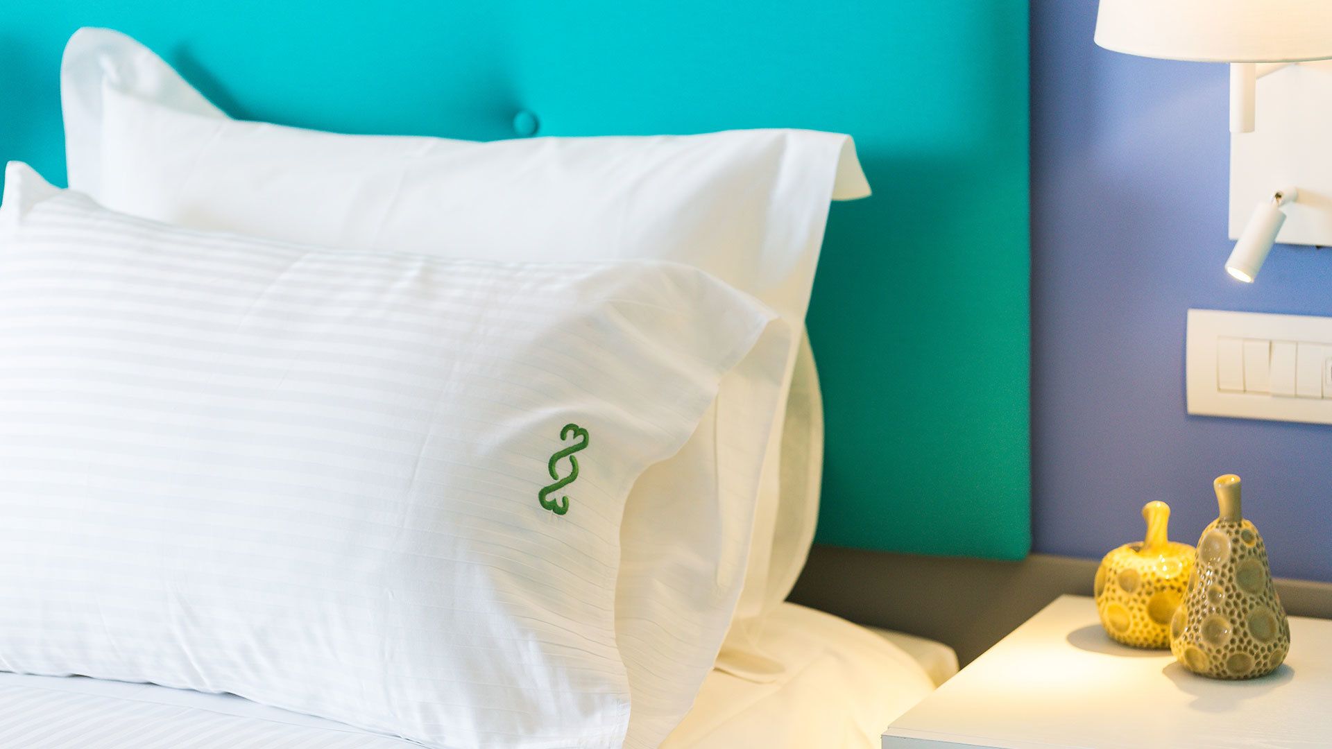A neatly made bed with white linens and a green embroidered pillow. There is a nightstand with two decorative vases and a wall socket.