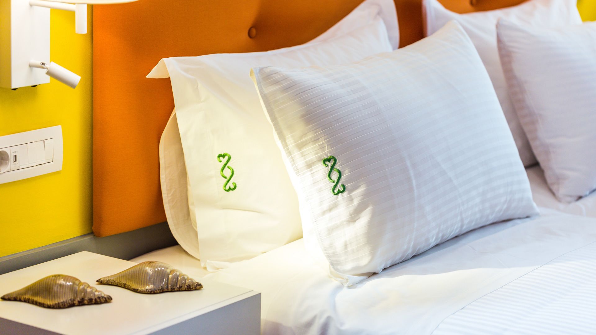 A bed with white pillows, a nightstand with a shell, and a wall socket.