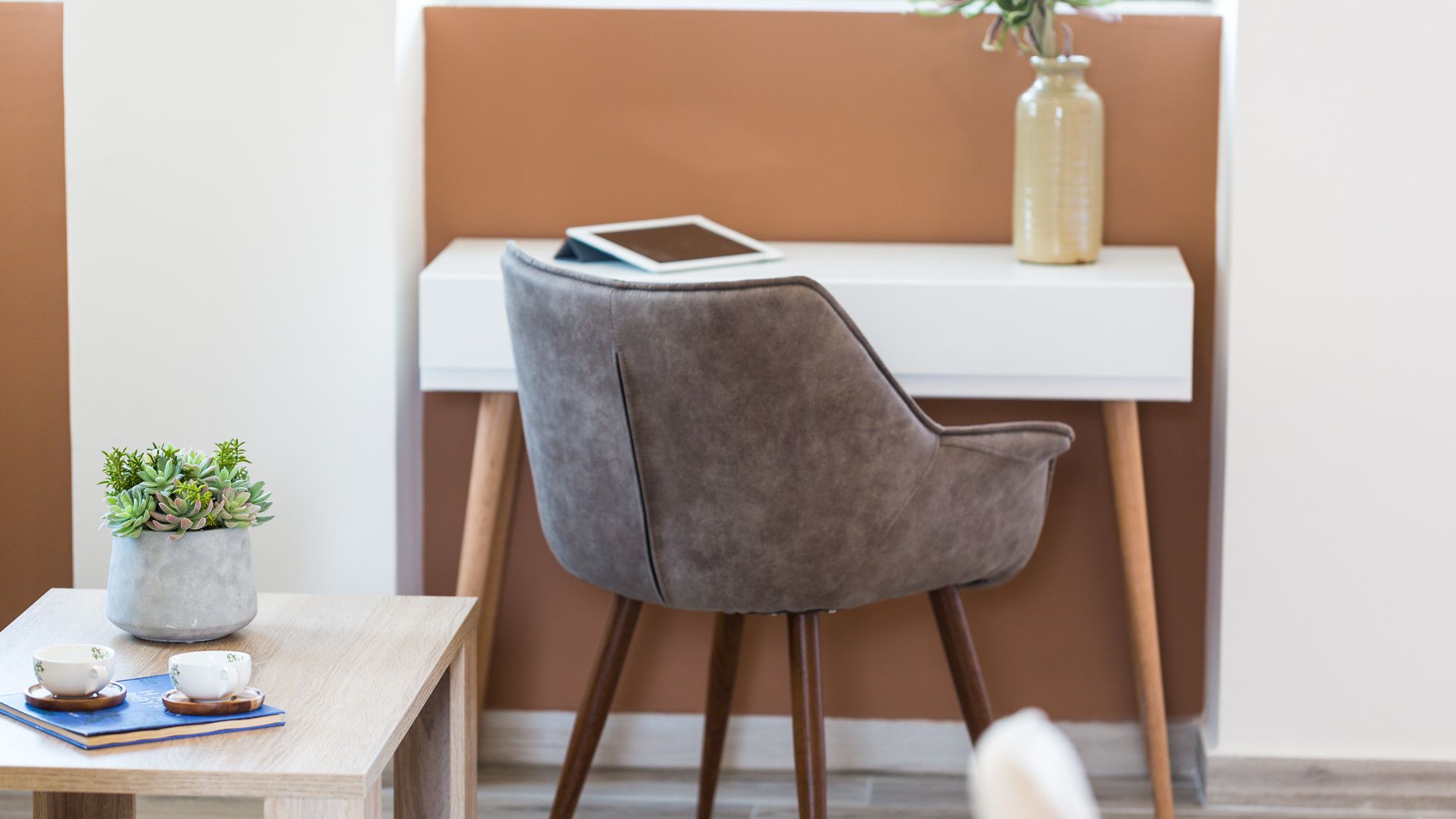 A modern room with a grey upholstered chair, a small table with a plant, a tablet, and a vase.
