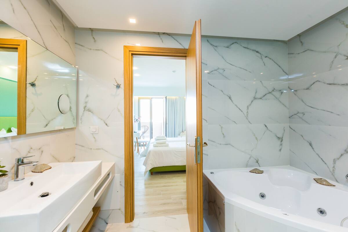 Un baño moderno con un espejo grande, dos lavamanos, una bañera y una puerta de madera que da a un dormitorio.