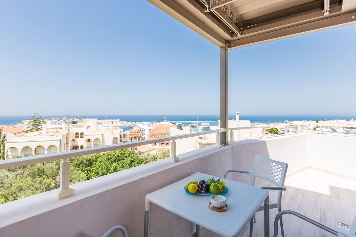Un balcón con mesa y sillas, un frutero y vistas al mar y al paisaje urbano.