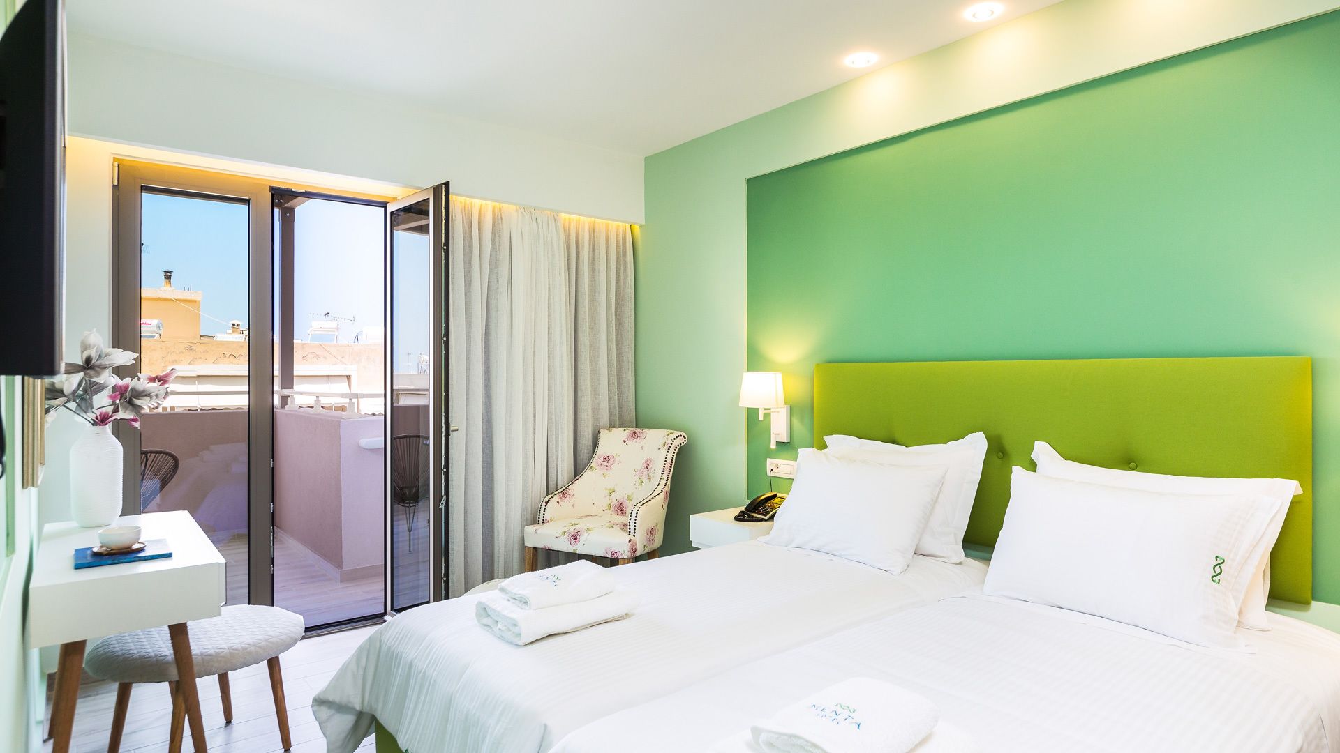 A hotel room with a green accent wall, a bed with white linens, a chair, a desk with a vase of flowers, and a balcony with a view of buildings.