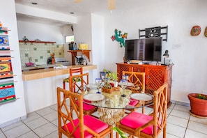A dining area with a round glass table, wicker chairs with pink cushions, and a TV mounted on the wall.