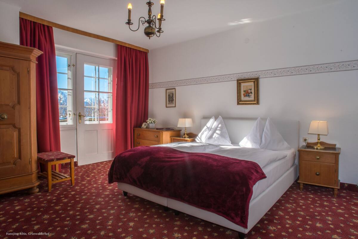 A bedroom with a large bed, two bedside tables with lamps, a wooden wardrobe, a small stool, and a door with red curtains.