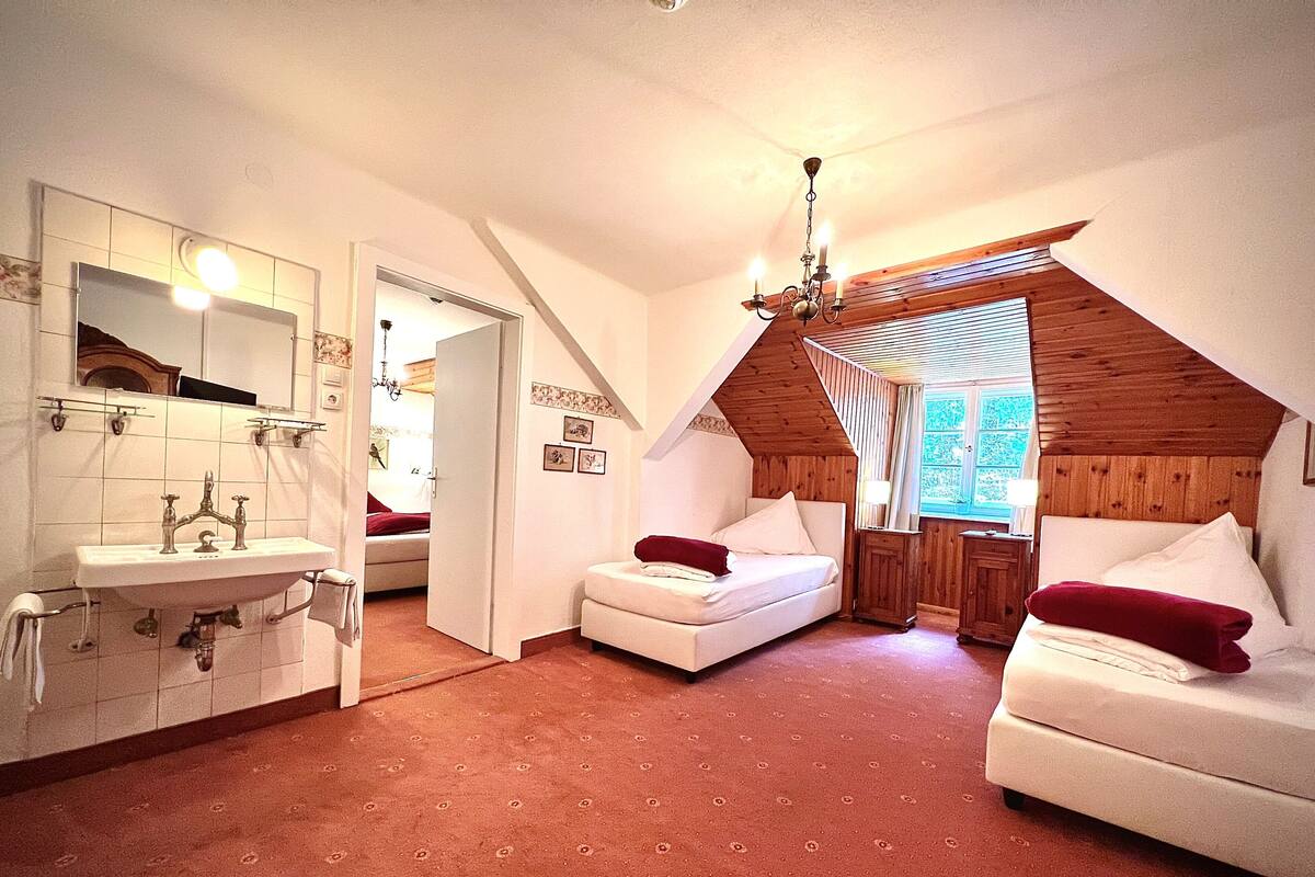 A bedroom with a sloped ceiling, a white bed, a white sofa, and a bathroom with a sink and mirror.