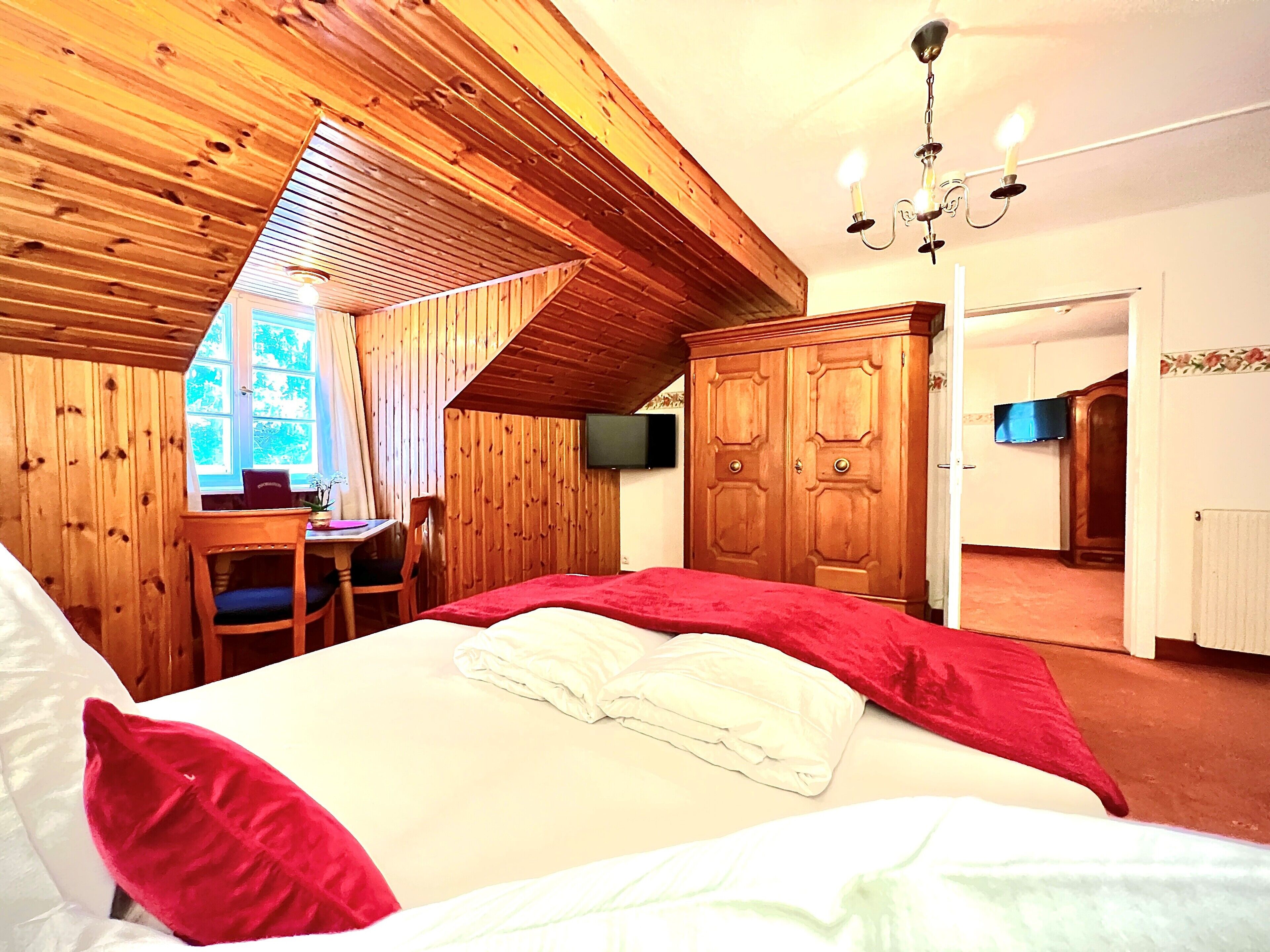 Un dormitorio con techo inclinado, paredes de madera, cama con ropa de cama roja y blanca, escritorio con silla y armario.