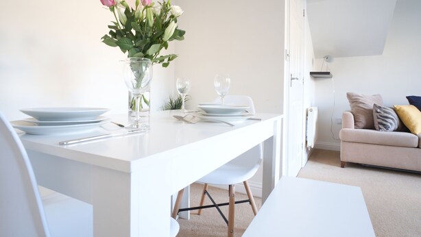 Un espace de repas moderne avec une table blanche dressée pour deux personnes, un vase rempli de fleurs et un canapé en arrière-plan.