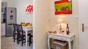 A kitchen with a refrigerator, dining table, chairs, and a small white cabinet with a vase and a painting. - Loggos Panoramic Views Electra By Konnect (Paxos)
