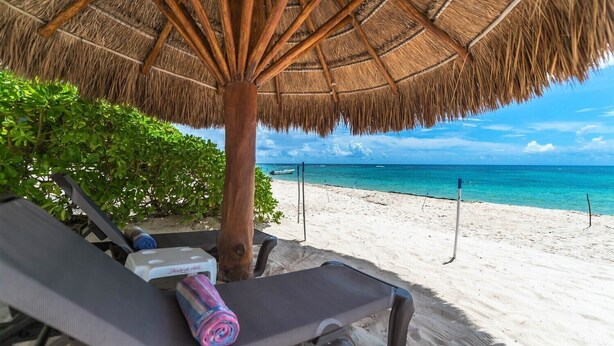 Un espacio de relax frente a la playa con una sombrilla de paja, vistas al mar y una tumbona con una toalla enrollada.