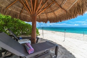 A beachside lounge with a thatched umbrella, a beach view, and a sun lounger with a rolled-up towel.