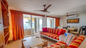 Un salón amplio con un sofá grande, una mesa de centro y una mesa de comedor. Hay ventiladores de techo, un ventanal con cortinas y vista al océano.
