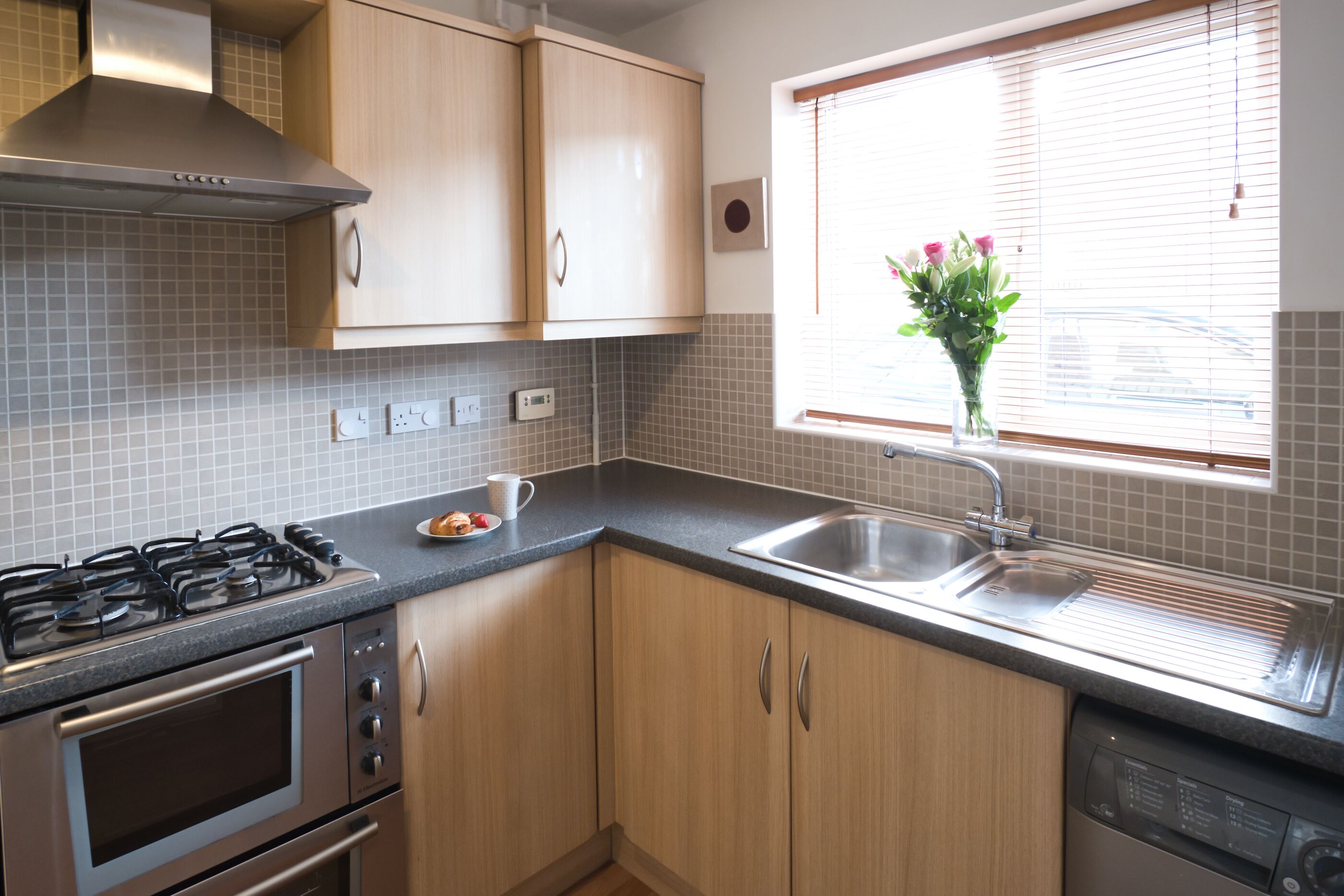 A modern kitchen with stainless steel appliances, wooden cabinets, and a window with blinds.