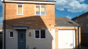 A two-story brick house with a front door, two windows, a garage door, and a small garden area with a potted plant. - KVM - Peacock Apartment (Peterborough)