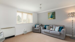 A living room with a grey sofa, a white radiator, a window with blinds, a floor lamp, and a painting of a peacock. - KVM - Peacock Apartment (Peterborough)