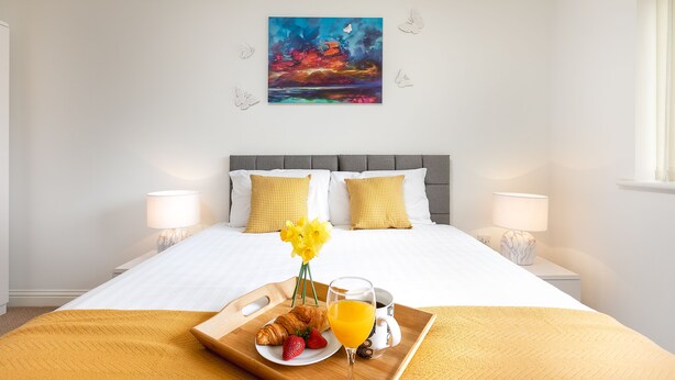 A hotel room with a bed, two bedside lamps, a painting on the wall, and a breakfast tray with croissants, strawberries, and orange juice. - KVM - Peacock Apartment (Peterborough)