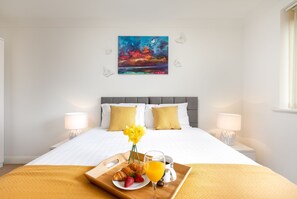 A hotel room with a bed, two bedside lamps, a painting on the wall, and a breakfast tray with croissants, strawberries, and orange juice.