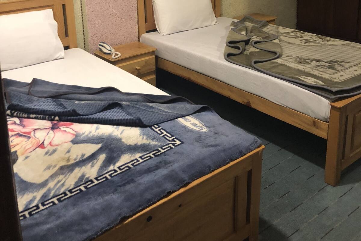 Ein kleines Zimmer mit zwei hölzernen Betten, jeweils mit einem weißen Kopfkissen und einem Holzkopfstück. Am linken Bett steht ein Nachttisch mit einer Lampe und einem Telefon. Eine blau gestreifte Decke mit Blumenmuster liegt über dem linken Bett drapiert.