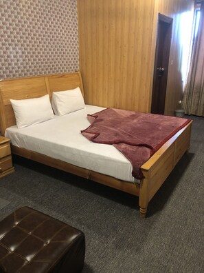 A wooden bed with white linens and a red blanket, a brown leather ottoman, and a patterned wall. - Rose Palace Hotel (Islamabad)