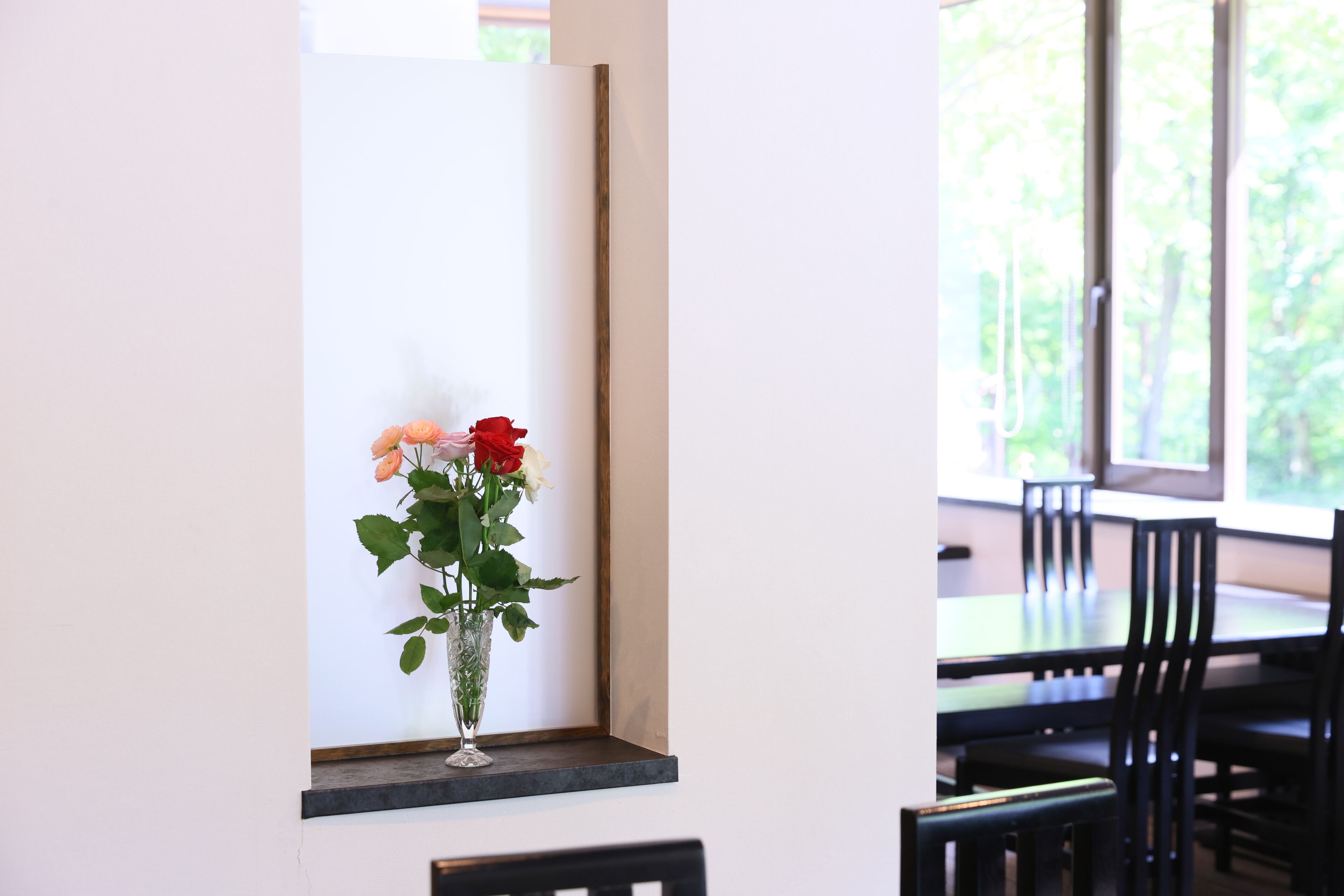 A dining area with a table and chairs, a vase with flowers on a shelf, and a window with a view of greenery.