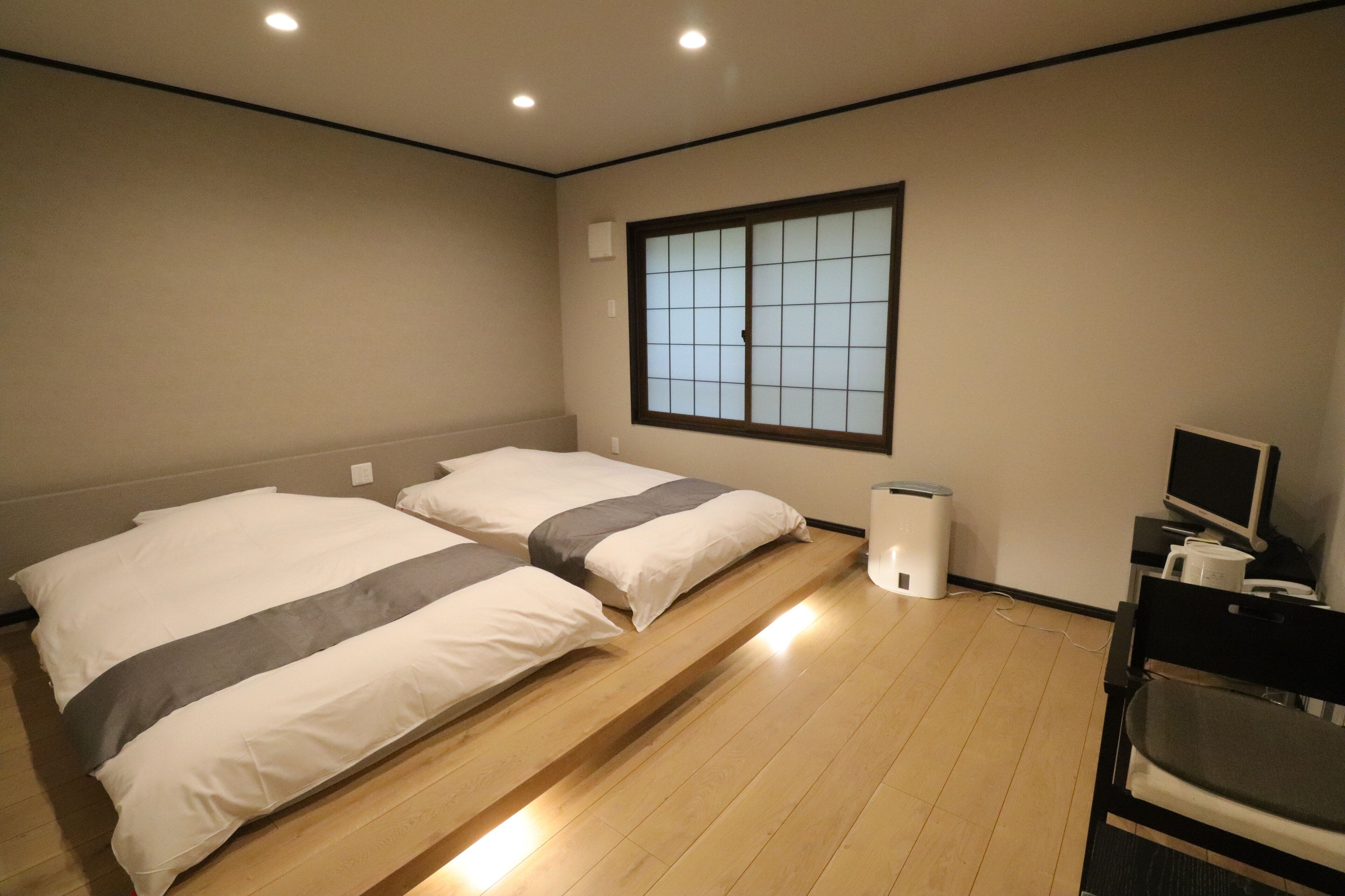 Ein Zimmer mit zwei Betten, einem Fenster mit Gittermuster, einem Schreibtisch mit Computer und einem Mülleimer.
